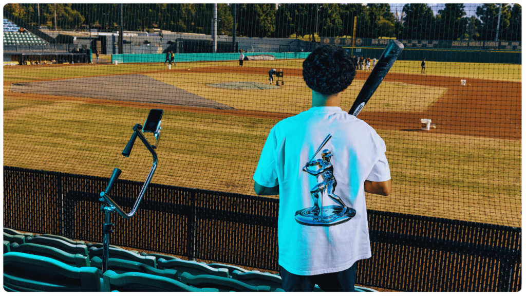 Teen baseball player holding a bat and watching the field from the stands, with an Attack Tee and phone setup nearby — ideal baseball tee for 14-year-old players to enhance hitting skills through video analysis.