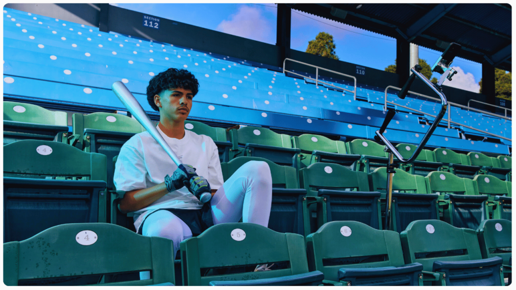 High school baseball player sitting in stadium seats with bat and batting tee, preparing for training with the best batting tee for high school players.