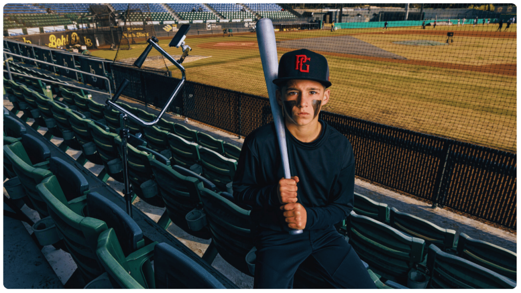 Young baseball player sitting in stadium stands holding a bat, with the Attack Tee—best baseball tee for 10-year-old players—set up near the field for batting practice.