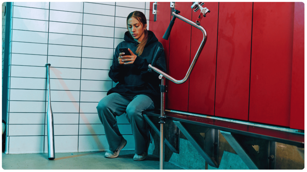 Female high school baseball player in locker room sitting beside the best batting tee for high school players, holding a phone before training.